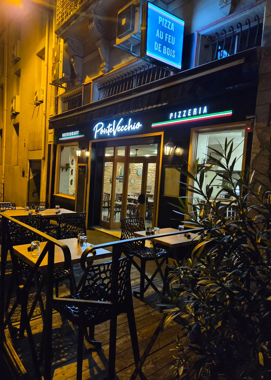 Façade du Ponte Vecchio de nuit : enseigne lumineuse « Ponte Vecchio Pizzeria », panneau « Pizza au feu de bois » et terrasse dressée