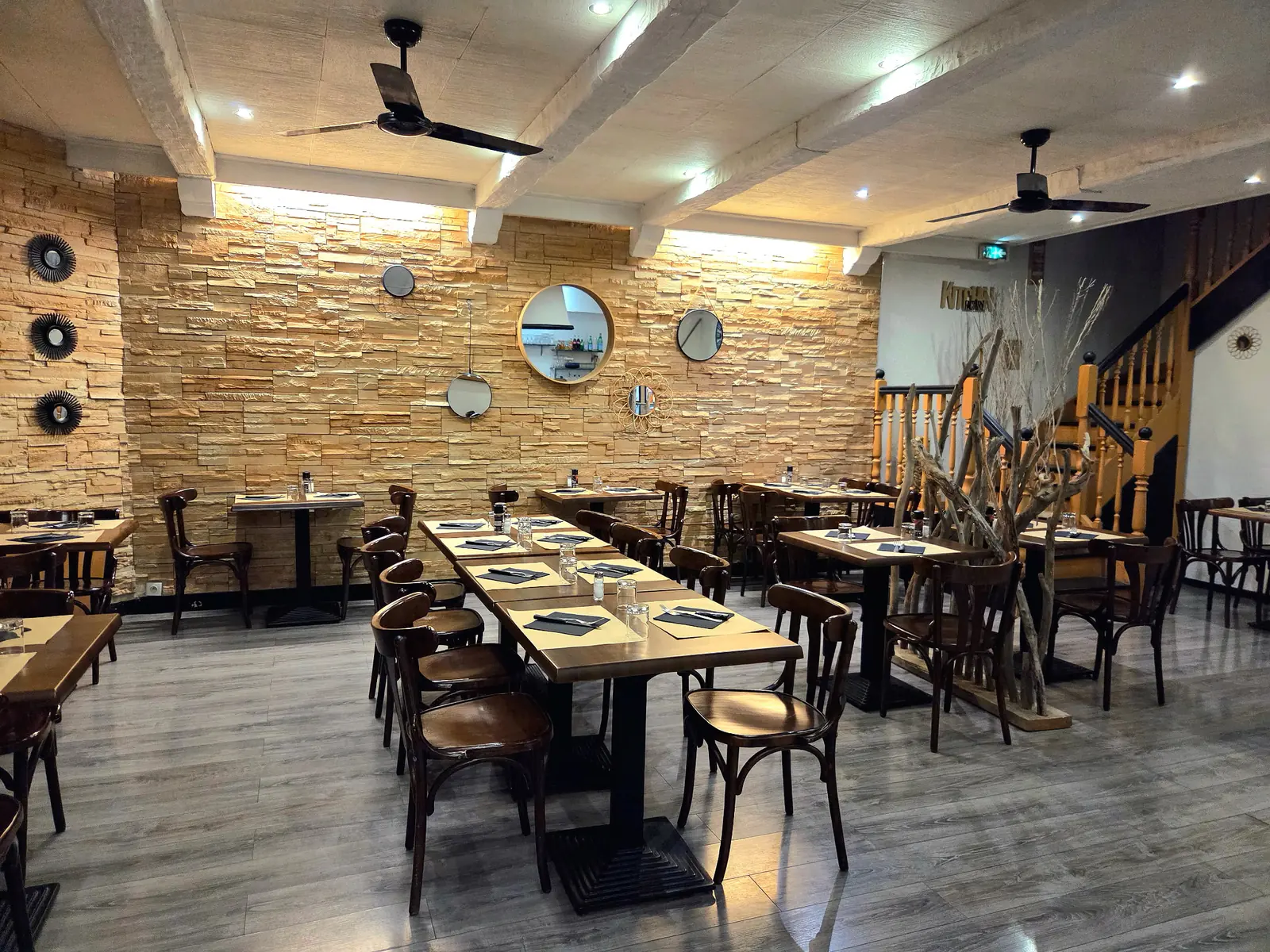 Salle du restaurant : mur en pierre éclairé, miroirs ronds, tables en bois dressées et ventilateurs au plafond